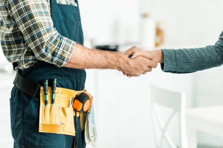 Plumber in a plaid shirt, navy overalls, and tool belt shakes hands with a customer inside a bright home.