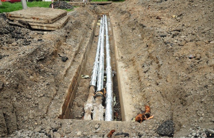 Excavated trench shows sewer line replacement with exposed pipes, wrapped sections, and soil piles at a plumbing project site.