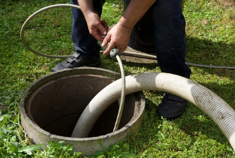 Worker wearing dark blue pants and black work shoes is cleaning a sewer drain by feeding a hose into an open concrete access point on a grassy lawn.