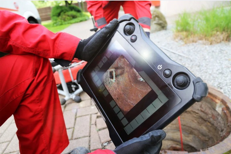 Technician in red coveralls and black gloves uses a sewer camera monitor to inspect pipe damage near an open outdoor drain.