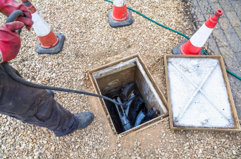Worker in muddy boots and red gloves guides an inspection hose into an open sewer drain beside safety cones.